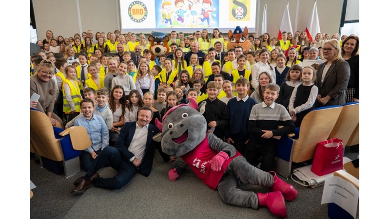 Grupowe zdjęcie dzieci i dorosłych w sali. Na pierwszym planie leży maskotka w czerwonej koszulce, a w tle widać uśmiechniętych uczestników, inne maskotki, ekran z grafiką oraz biało-czerwone flagi fot. FB szkoły